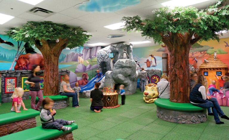 Zoo-themed kid's play area with custom tree bench seats, colorful wall murals, sculpted rock activity stations, play structure and soft-play climbable characters.