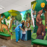 A dentist and young girl stand at a sink in a brightly lit dental clinic, showcasing a themed environment transformation with colorful murals of forest animals—bears, deer, and birds—creating a cheerful, child-friendly atmosphere.