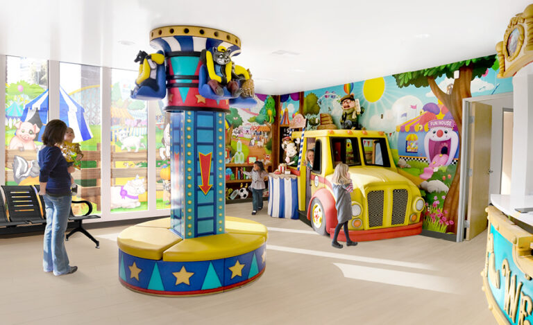 Brightly colored indoor playroom with carnival themed decor, including cartoon murals, a yellow play area food truck, a seating bench, and a spinning ride featuring animal figures. Several children are playing while an adult stands nearby. Large windows let in natural light.