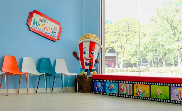 A colorful waiting area with orange, teal, and white chairs, a large window, and a smiling popcorn mascot. The walls are blue with a My Smiles sign, and a bench decorated with film and cartoon popcorn images.