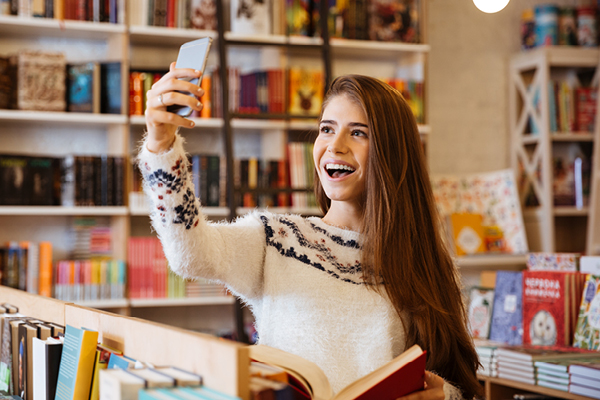 A young woman with long brown hair smiles and takes a selfie with her smartphone while holding an open book in a brightly lit bookstore filled with colorful books.