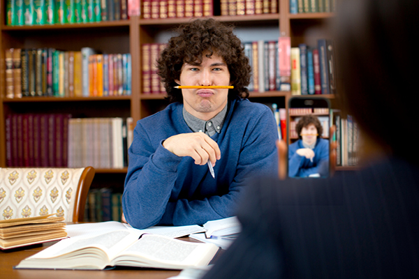 A young man with curly hair balances a pencil on his upper lip while sitting at a table with open books, looking playfully at a camera phone across from him in a library setting.