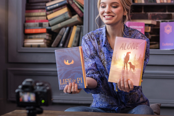 A smiling woman sits indoors holding up two books, Life of Pi and Alone, in front of a camera. Bookshelves filled with colorful books are in the background.