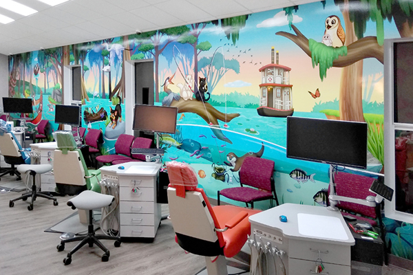 A colorful medical room with several exam chairs and monitors features a vibrant wall mural of a river scene with trees, animals, and a steamboat, creating a cheerful, child-friendly environment inspired by an IDS testimonial.