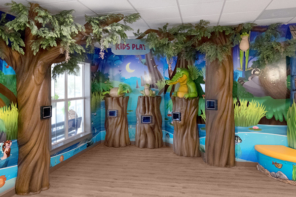 A colorful indoor kids’ play area with tree decorations, animal figures, interactive screens on tree stumps, and a mural of a forest and pond on the walls. An IDS testimonial highlights its inviting atmosphere. The sign reads KIDS PLAY; wood-patterned flooring adds charm.