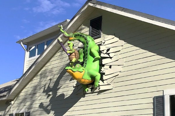 A large sculpture of a turtle riding an alligator appears to burst through the exterior wall of a beige house, with broken siding around them, like an IDS testimonial brought to life. The sky is clear and blue in the background.