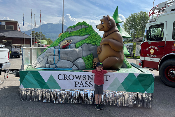 A young boy stands in front of a parade float adorned with a large bear, rocks, and trees under a Crowsnest Pass banner. A fire truck is parked beside the colorful parade float on a sunny day.