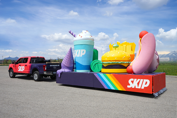 A red pickup truck with SKIP branding tows one of the parade floats, featuring oversized models of a drink, ice cream cone, fries, burger, and hot dog on a rainbow platform, all set outdoors under a blue sky.