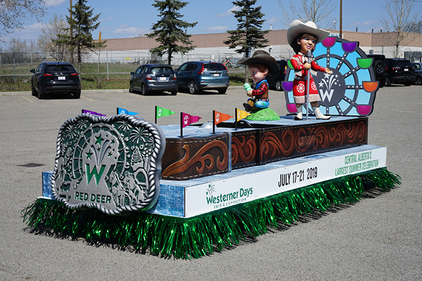 Parade floats with a “Red Deer Westerner Days” theme feature cowboy and cowgirl figures, colorful signs, and event dates July 17-21, 2019. The vibrant float is parked in a lot with trees and cars in the background.