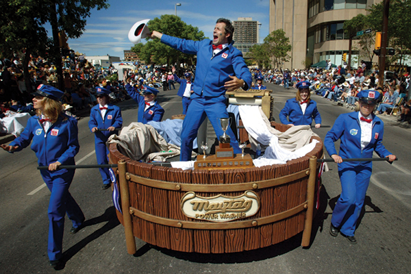 A group of people in blue uniforms pull a large wooden parade float labeled Maytag Power Washer during a parade. One person stands on the float, raising a hat, while crowds watch from the sidewalks.