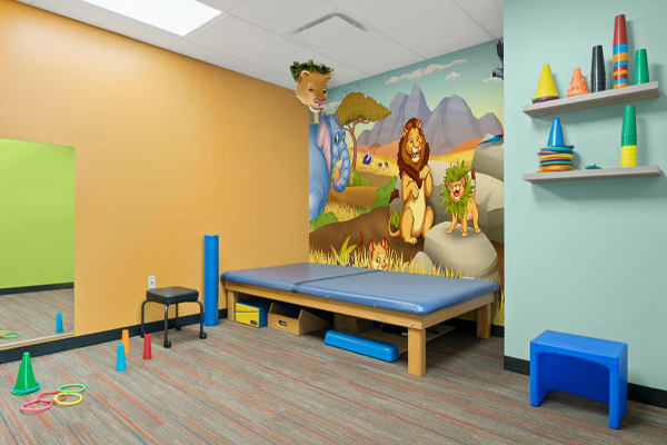 A cheerful pediatric therapy room showcases vibrant therapy clinic decor with colorful cones, rings, and a blue bench. A padded therapy table rests against a wall featuring a mural of cartoon jungle animals like a lion and giraffe.