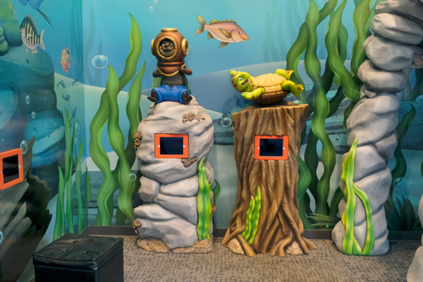 An underwater-themed play area with tall rocks, a sea turtle on a tree stump, a diver helmet, colorful fish, and seaweed decorations. Two screens are embedded in the rocks.