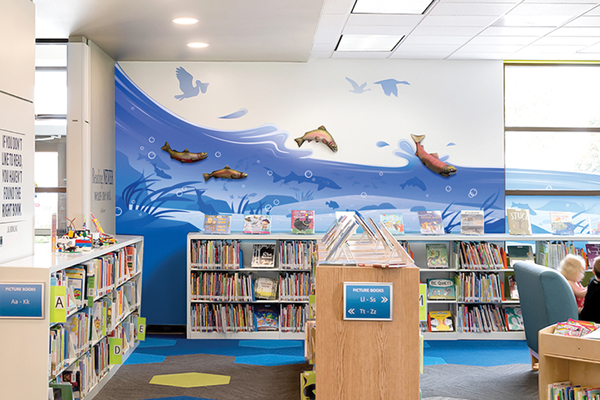 A colorful childrens library with low bookshelves, bins filled with picture books, and a blue wall mural featuring fish, birds, and waves. Children are reading in a cozy corner near large windows.