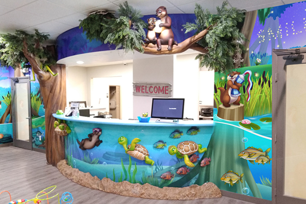 A colorful reception desk decorated with cartoon animals, including turtles, otters, and birds, under a tree with playful characters and underwater scenes. A “WELCOME” sign is visible behind the desk.