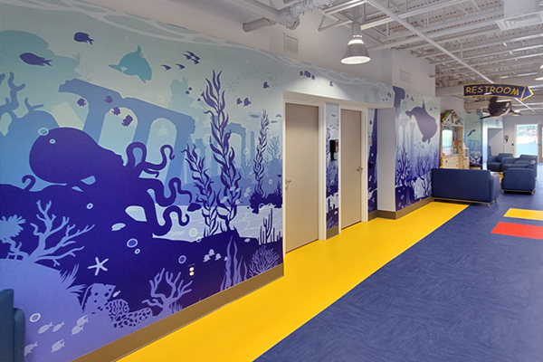 A hallway with yellow and blue floors features a vibrant underwater-themed mural with sea creatures, plants, and ruins. There are multiple doors and a restroom sign, with blue couches in the seating area.