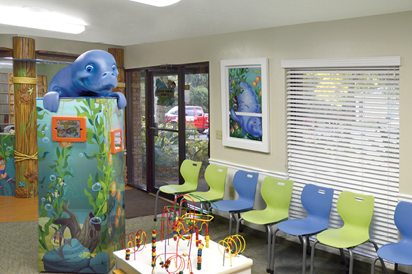 A bright pediatric waiting room with blue and green chairs, a play table with bead mazes, colorful aquatic-themed wall art, and a large sculpted manatee figure on a video game tower.