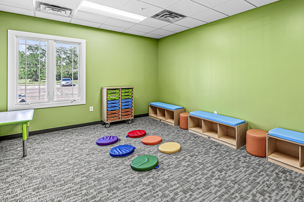 A bright, green-walled classroom with a window, blue benches, storage cubbies, and colorful round floor cushions arranged in a circle on gray carpet. A cart with green trays stands near the wall.
