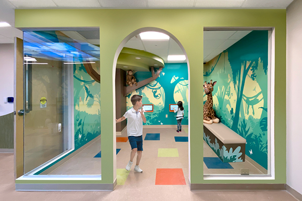 A brightly colored pediatric playroom with jungle-themed decor features wall murals, two kids playing, a curved slide, a plush giraffe, and colorful square tiles. Large windows and an archway frame this cheerful hospital space.