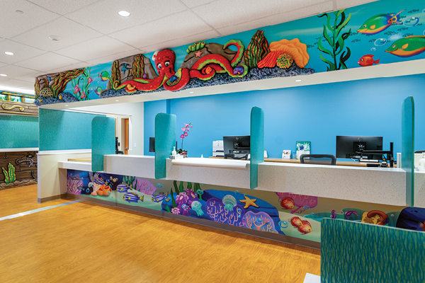 A brightly colored hospital reception area features themed decor with an underwater motif, including murals of sea creatures like an octopus and fish on walls and counters, plus aqua blue accents throughout the medical space.