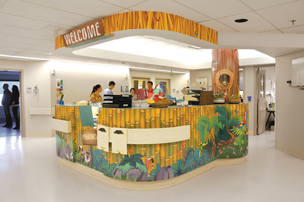 A hospital reception desk adorned with colorful jungle-themed decor and a “WELCOME” sign, where medical staff assist people behind the counter in a brightly lit hallway.