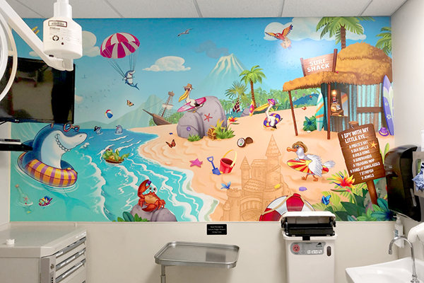 A colorful pediatric mural of a tropical beach with cartoon animals surfing, building sandcastles, and relaxing near water brightens a medical exam room, featuring themed decor with equipment and a sink in the foreground.
