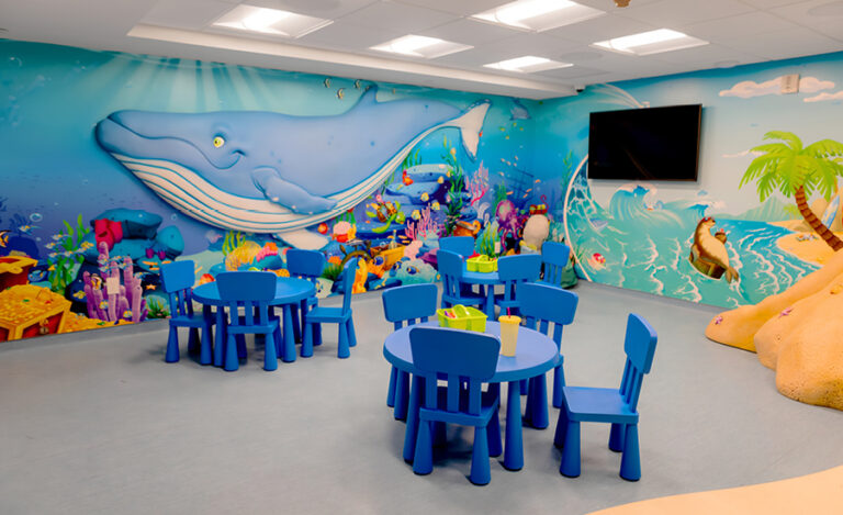 Colorful classroom for kids ministry with blue child-sized tables and chairs, ocean-themed wall mural featuring a large whale, fish, and sea scenery, and a television on the wall. The room is bright and playful—perfect for a church setting.