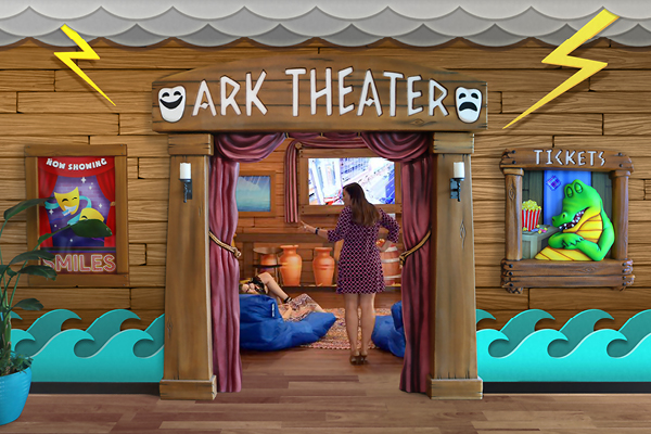 A woman stands at the entrance of a whimsical, ocean-themed Noah's Ark themed theater with cartoon waves and scenic fabrication details. Inside this playful themed environment, two children lounge on blue bean bags, watching a screen.