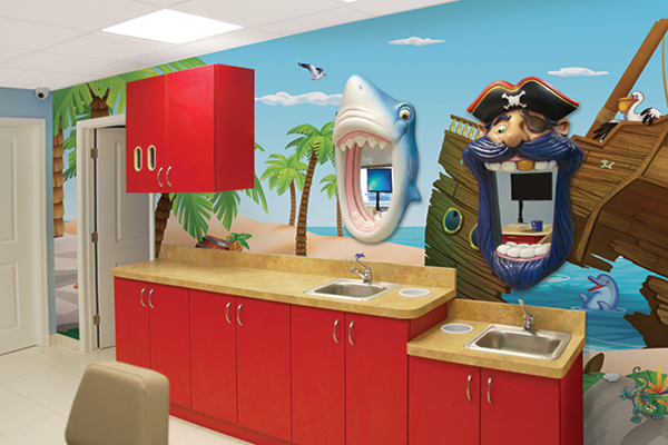 A brightly lit dental office with red cabinets and playful themed decor, including a cartoon shark and pirate with wide open mouths, palm trees, a shipwreck, and a beach scene mural that creates a fun, imaginative environment for kids.