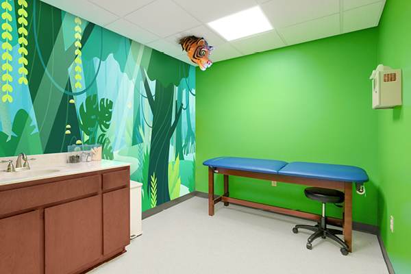 A brightly lit medical exam room with green walls, a jungle-themed mural, a blue exam table, a brown cabinet with a sink, and a cartoon tiger head decoration hanging from the ceiling.