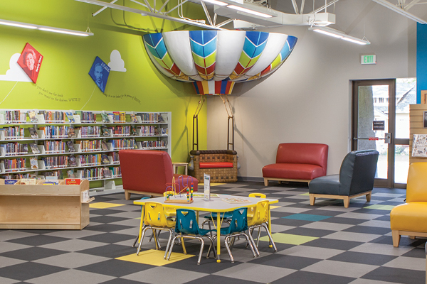 This themed environment features a colorful children's library area with bookshelves, small tables and chairs, cushioned seating, and a decorative hot air balloon display. The bright green walls are adorned with colorful kites, creating playful scenic fabrication.