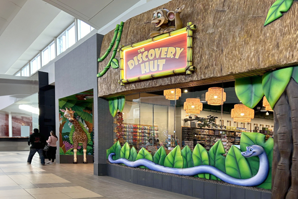 A colorful store called The Discovery Hut features a jungle-themed store facade with a retail sign, a giraffe, smiling snake, and cheerful animal decorations. Two people walk past in the mall’s bright, spacious corridor.