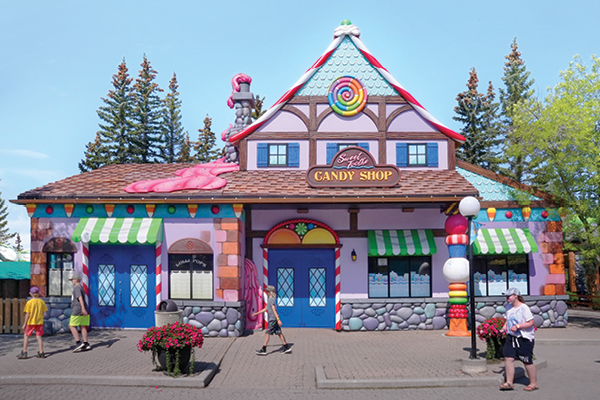 A whimsical candy store with colorful decorations, including candy-shaped sculptures on the roof and candy cane striped awnings. People walk by the playful, pastel-painted building in this charming themed environment on a sunny day.