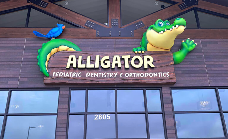 Sign for Alligator Pediatric Dentistry & Orthodontics with a cartoon alligator and blue bird on a wooden-style board above glass doors.