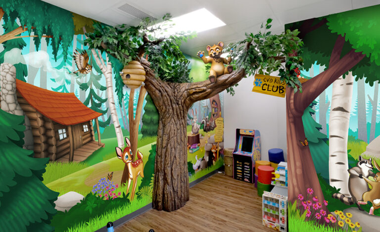 A colorful, cartoon-themed kids playroom in a pediatric dental office features a large artificial tree with leafy branches, animal murals on the walls, a beehive, play equipment, toys, and a CVO Kids Club sign hanging from the tree.