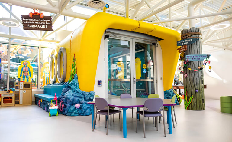 A colorful indoor play area features a yellow submarine structure with portholes, surrounded by beach-themed ocean decor, small tables and chairs, and playful signage, creating a cheerful, child-friendly environment.