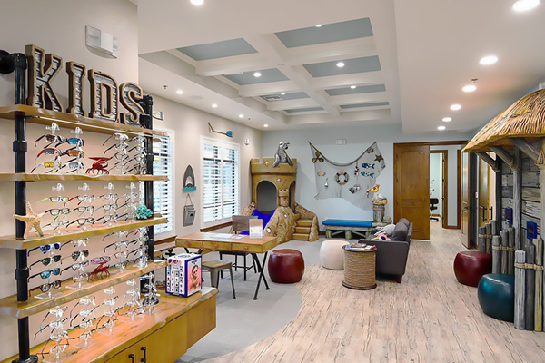 A bright and inviting children's optometry area featuring a "KIDS" sign, colorful frames display, and a beach-themed waiting space with ocean-inspired decor and seating shaped like marine animals.