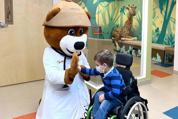 A child in a black and green wheelchair wearing a mask and a striped blue shirt high fiving a bear mascot in front of a giraffe sculpture and jungle themed mural inside of a pediatric unit in a hospital.