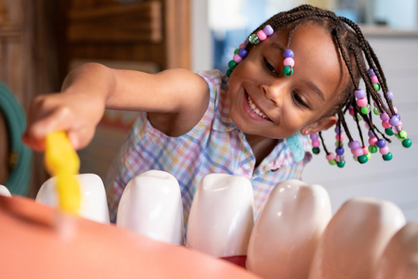 A child with multicolored beads and a plaid shirt using a yellow tooth brush to brush the teeth of a sculpted mouth including a sculpted tongue and sculpted teeth