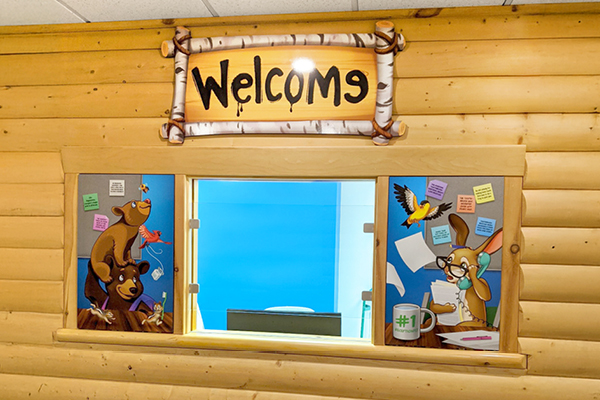 A wooden Pediatric Front Desk features a “Welcome” sign above a window, decorated with colorful cartoon animals like bears and a rabbit, plus sticky notes on either side to greet young visitors.