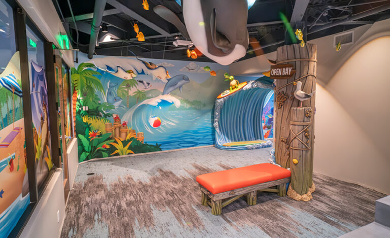 A colorful, beach-themed indoor play area in the dental office with ocean murals featuring dolphins, a red cushioned bench, a large wave slide, and playful sea decorations hanging from the ceiling.