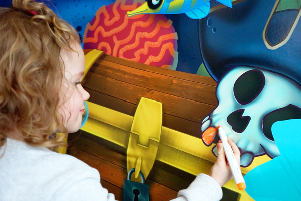 A young child with curly hair colors a cartoon skull on a treasure chest with an orange marker. Easy-to-clean IDS mural.