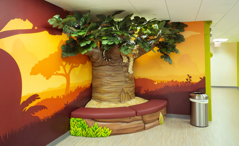 Safari themed decor in the waiting area of a pediatric medical facility. A whimsical indoor bench shaped like a tree trunk with leafy branches, set against a mural of a sunset landscape. The bench curves along the wall, accompanied by a silver trash can on light wood flooring.
