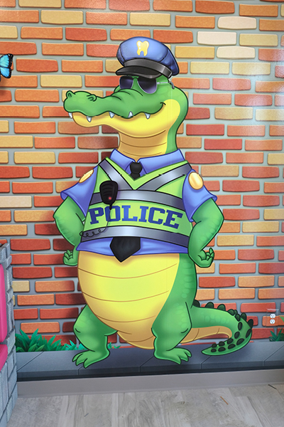 Cartoon alligator dressed as a police officer, wearing a blue uniform, hat, sunglasses, and a belt, standing confidently against a brick wall background.