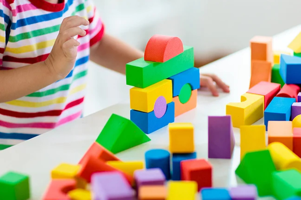 Waiting room toys and activities. A child wearing a colorful striped shirt is playing with bright, multicolored foam building blocks, stacking them on a white surface while more blocks are scattered around.