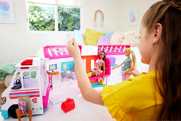 Waiting room toys and activities. A young girl with Barbie dolls and a colorful toy ambulance set in a bright, cozy room with a bed and pillows in the background.