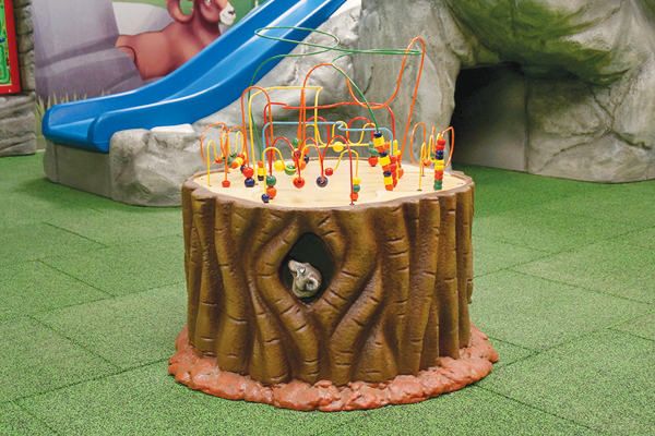 Waiting room toys and activities. A play area with a realistic tree stump activity table featuring bead maze wires, a meerkat figure peeking out of a hole, and a blue slide in the background on green padded flooring.