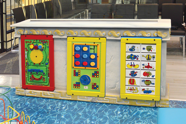 Waiting room toys and activities. A children's play panel with three colorful activity boards featuring gears, buttons, and animal-themed flip panels, set in an indoor play area with blue water ripple flooring.