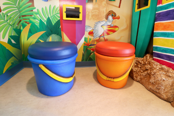 Two large, colorful stools shaped like beach pails—one blue with a yellow handle, one orange with a yellow handle—sit in a vibrant, tropical-themed room with a beach-themed wall mural.