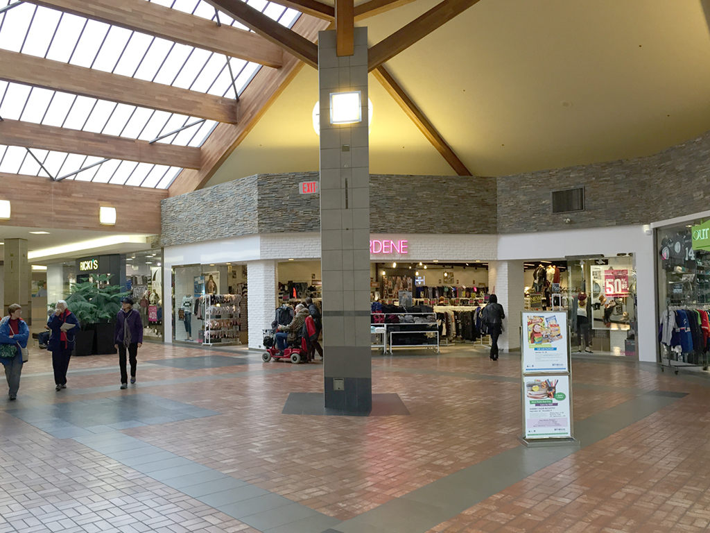 A drab mall interior with several people walking and shopping. Visible stores include Ardene, a shop with a “50% off” sign, and another with clothes on display. The ceiling is tall with skylights and wooden beams—this is a boring before photo.