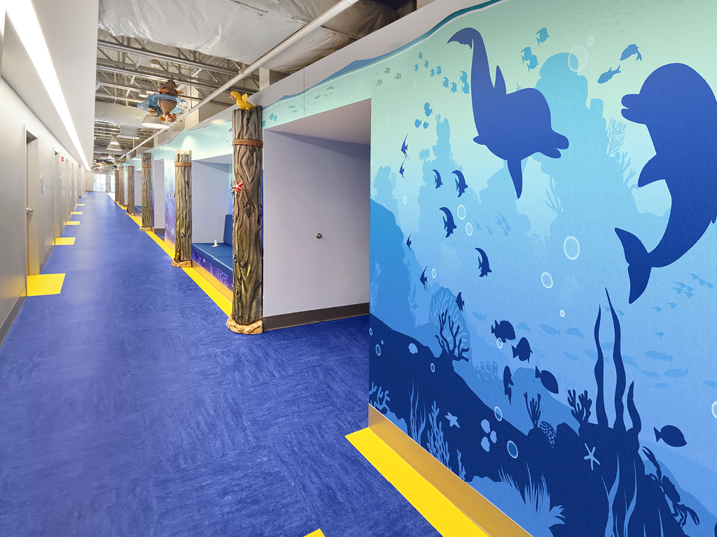 Experience a stunning themed environment transformation in this brightly lit hallway, where blue and yellow floors, wooden piling columns, and an underwater mural with dolphins create a vibrant before and after effect. Exposed ceiling pipes complete the look.